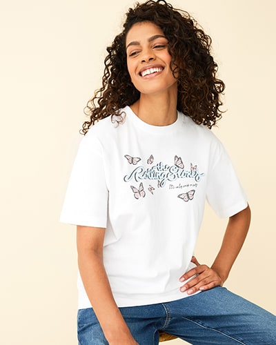 White T-shirt with the words the rolling stones written in light blue cursive script. Underneath are the words it's only rock 'n' roll. All words are surrounded by light pink butterflies.