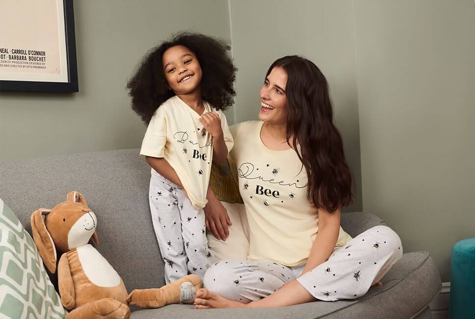 Pale yellow short sleeve top with 'Queen Bee' across the chest with small bees and hearts around it. Grey PJ bottoms with bee and heart print. Child is wearing a matching set with 'Bumble bee' written across the chest 
