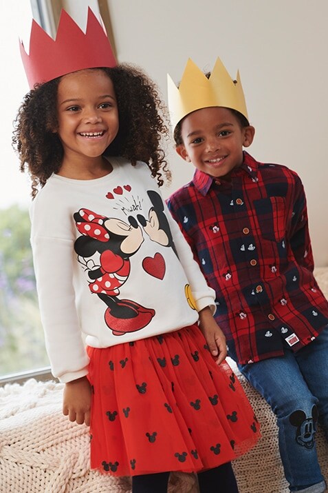  Girls' set features red skirt with black Mickey Mouse print, and a white, long-sleeved top with an image of Minnie and Mickey kissing. Boys' set features dark blue jeans with a black Mickey detail on the knees, and a red and blue checked shirt with Mickey mouse print.